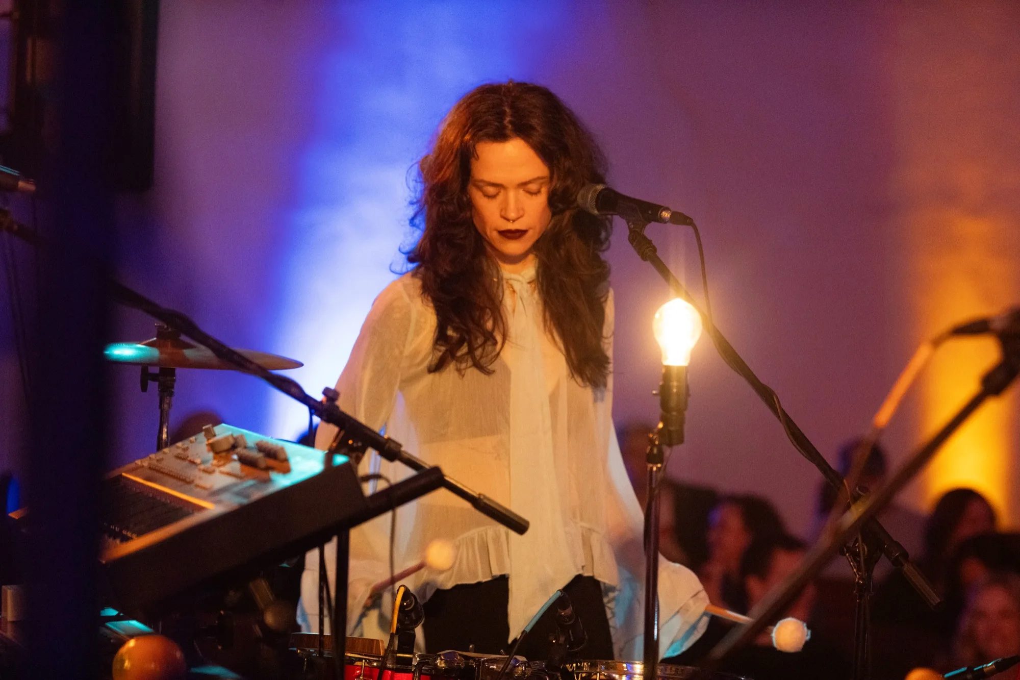 Amy Lee Montgomery singing into a mic with eyes closed, wearing a white ruffled top, playing drums, a keyboard just in view.