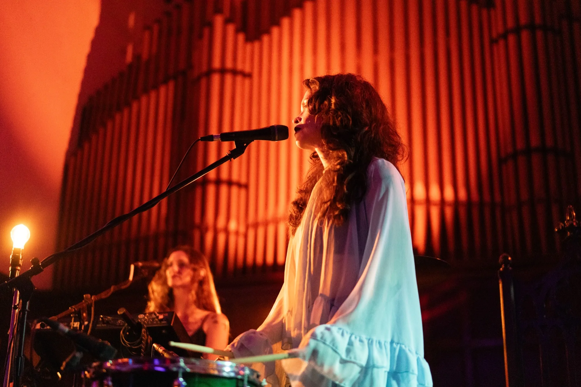 Amy Lee Montgomery in a white ruffled top, singing and playing drums in front of tall red-lit organ pipes.