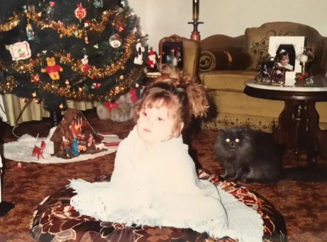 childhood photo of me sitting on a large pillow on the floor, a persian cat hissing behind me, next to a decorated christmas tree.