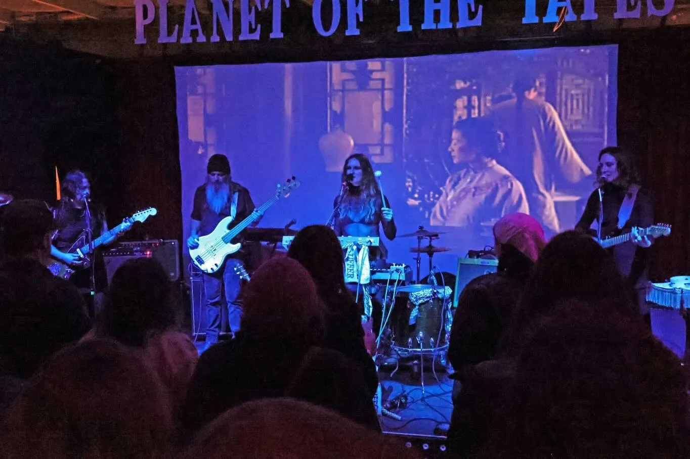 Band duchess performing on stage at Planet of the Tapes under blue and purple lights, with a film projected on a screen behind them.