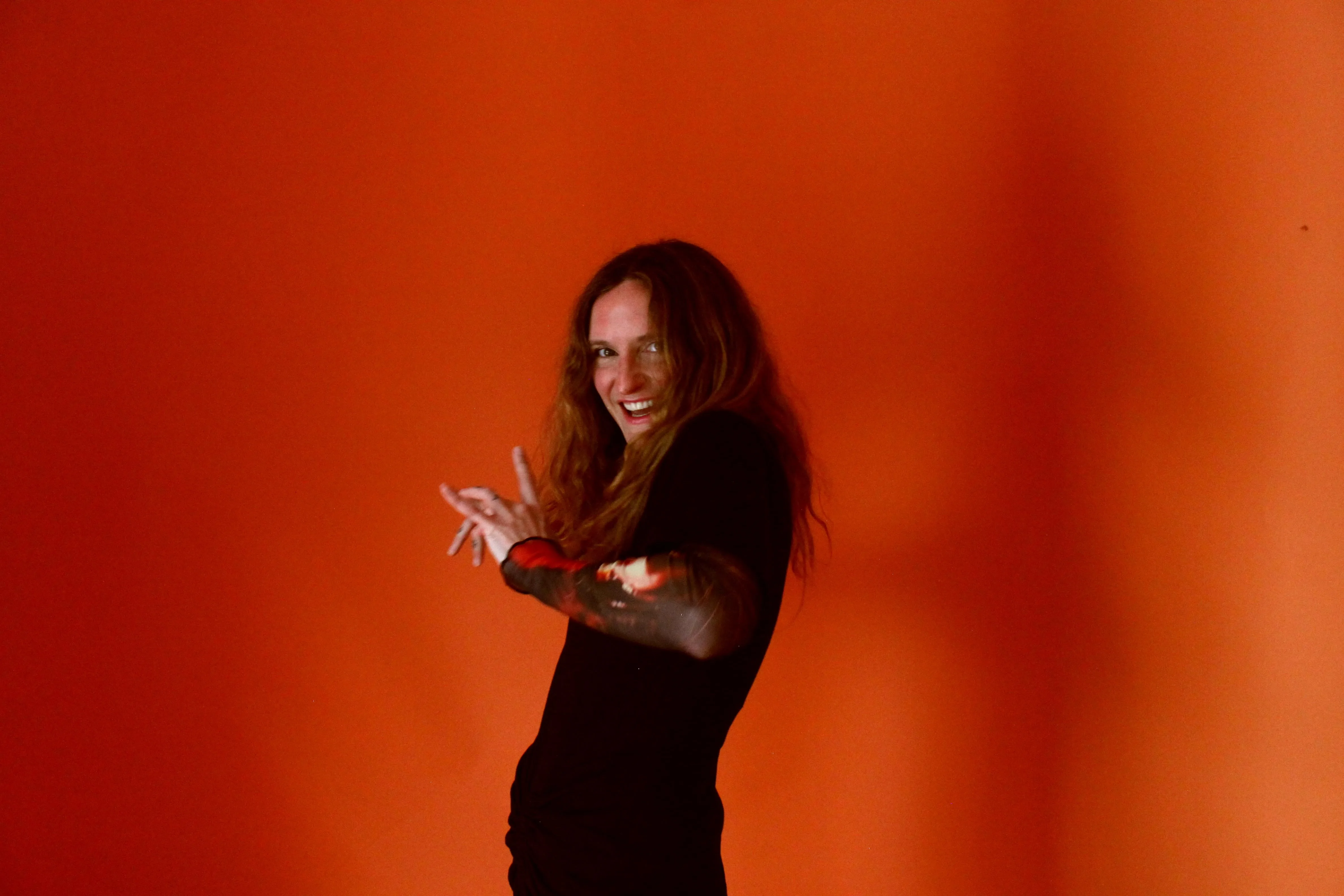Manic smile at the camera with my hands raised and curled in front of me against a deep orange backdrop.