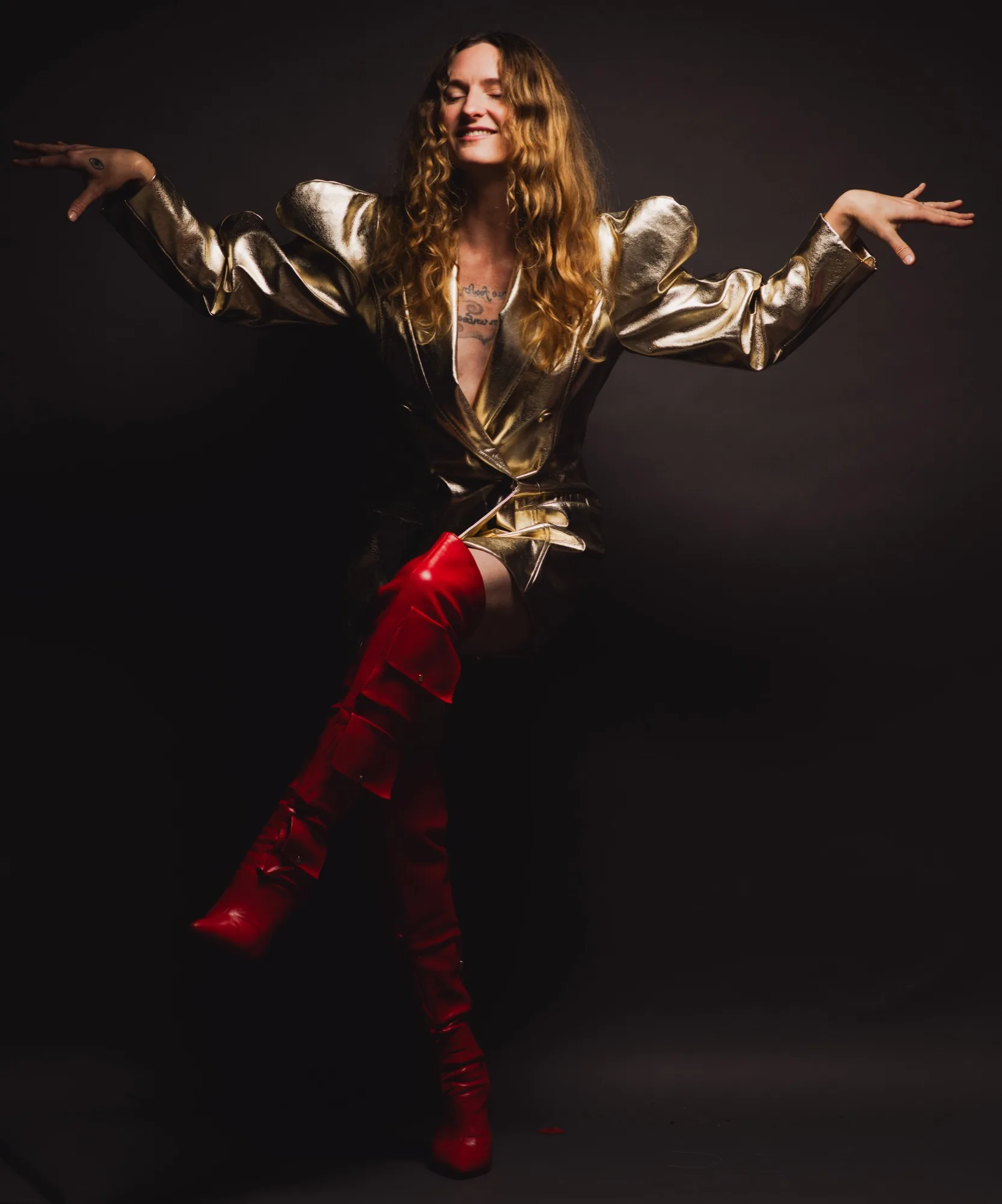 Lacey Guthrie balancing on one leg in a gold metallic blazer and red thigh-high boots, arms spread wide, eyes closed, grinning.