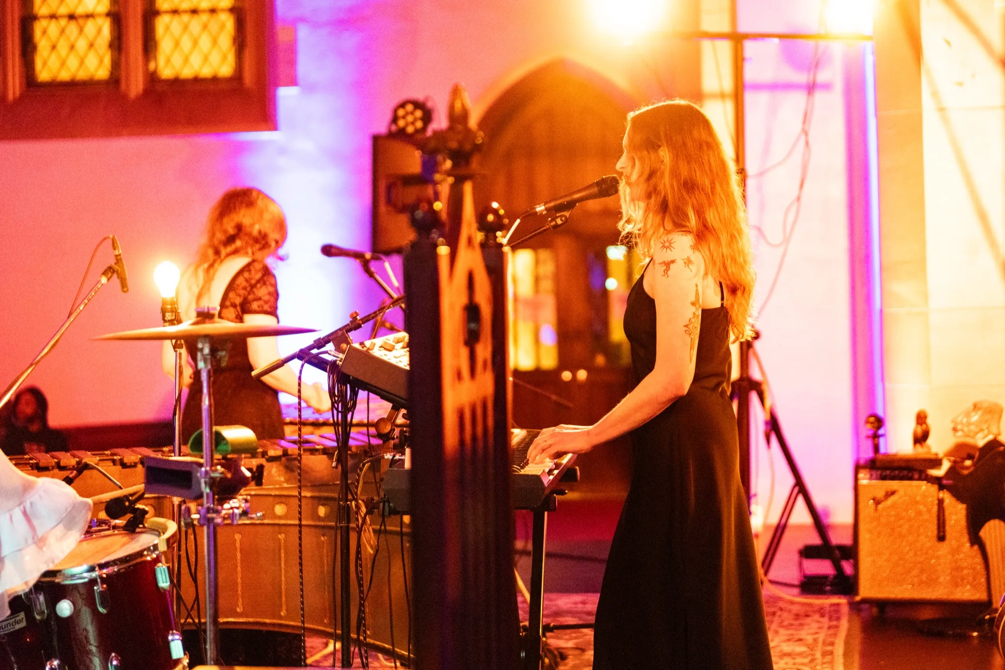 Lacey Guthrie singing at a keyboard while a bandmate plays drums behind her, under a stained glass window.