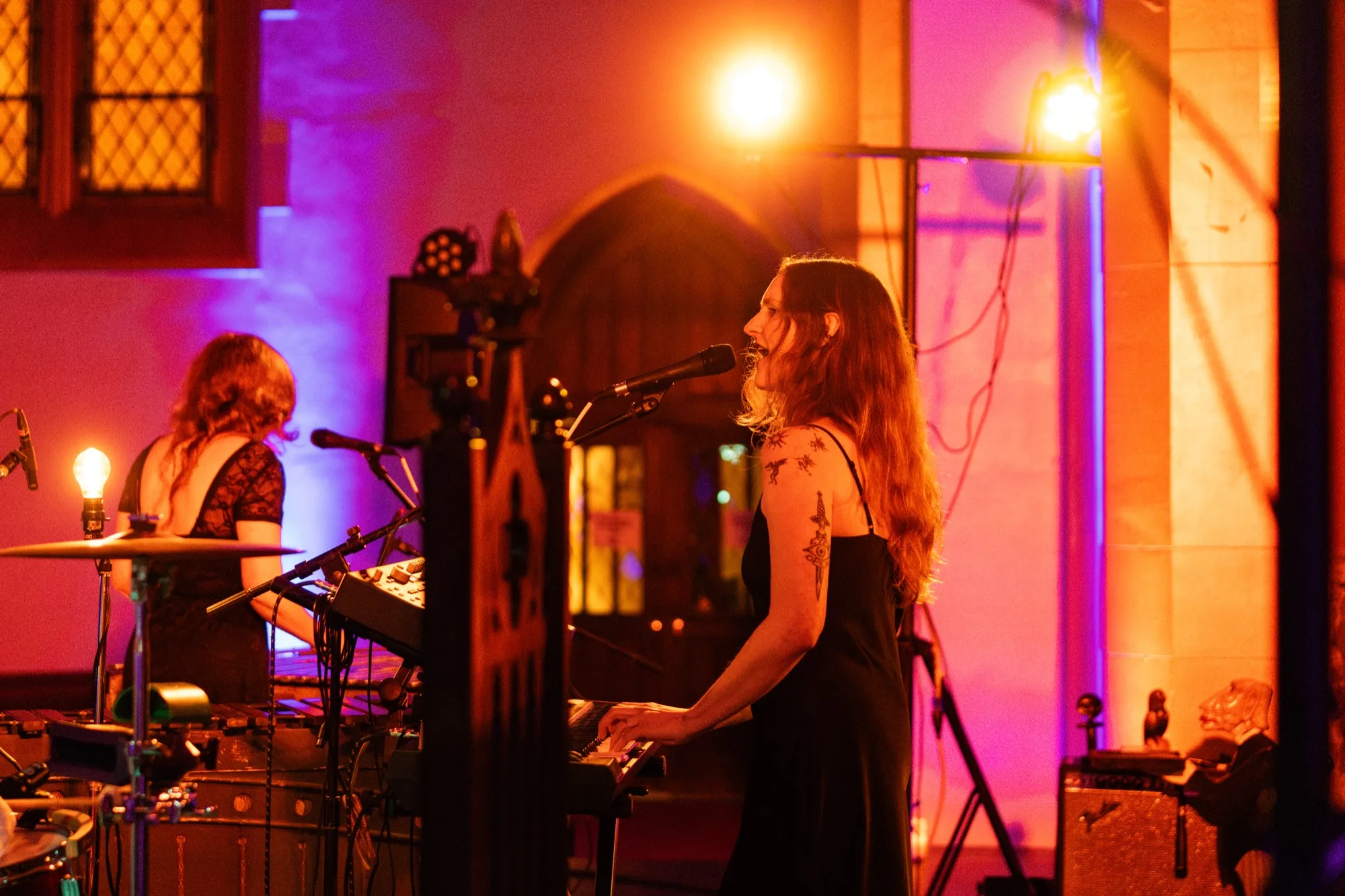 Lacey Guthrie singing at a keyboard in a black dress, a bandmate on drums behind her, under warm stage light.