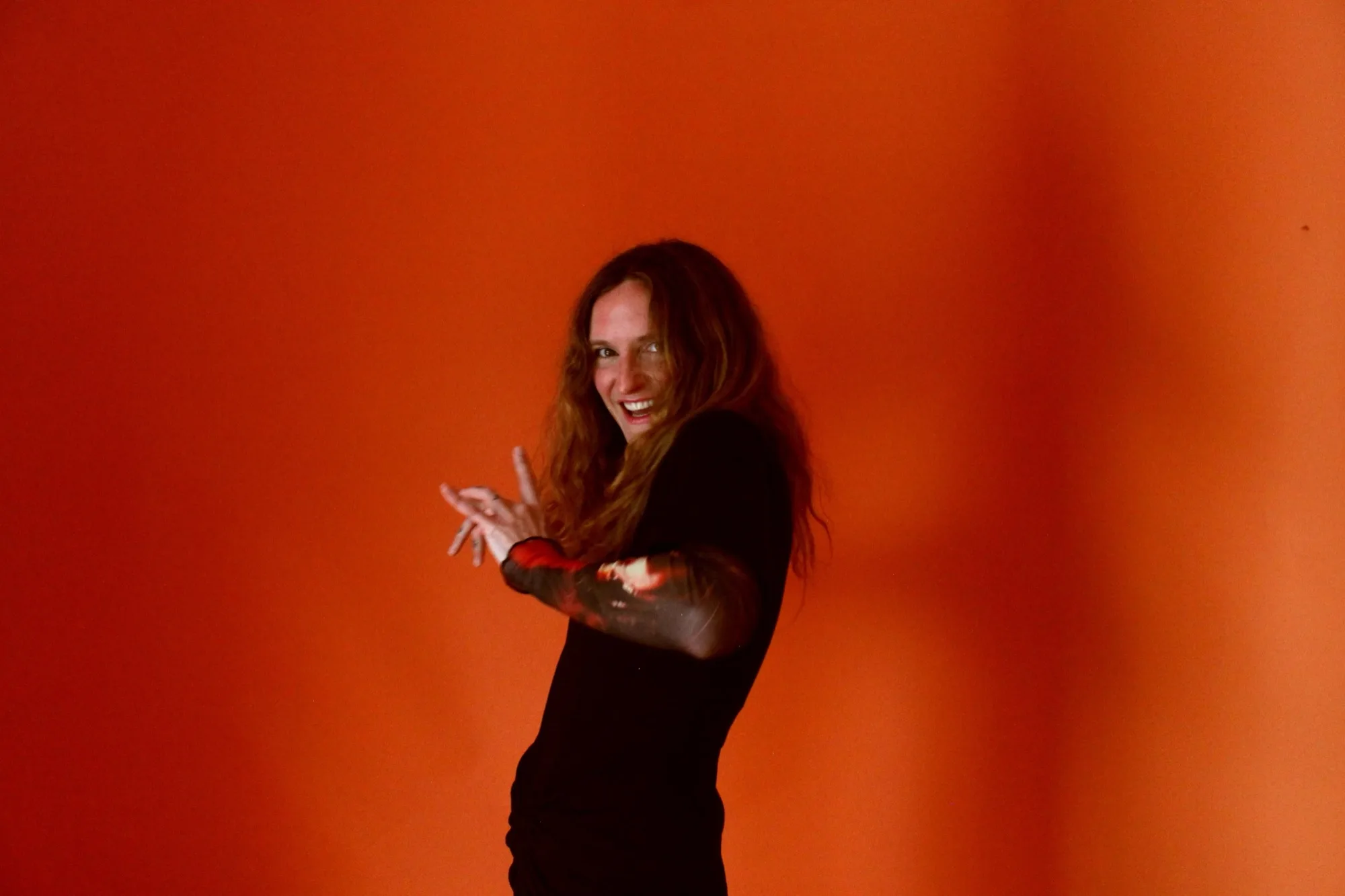 Lacey Guthrie smiling manically at the camera, hands raised and curled in front of her, against a deep orange backdrop.