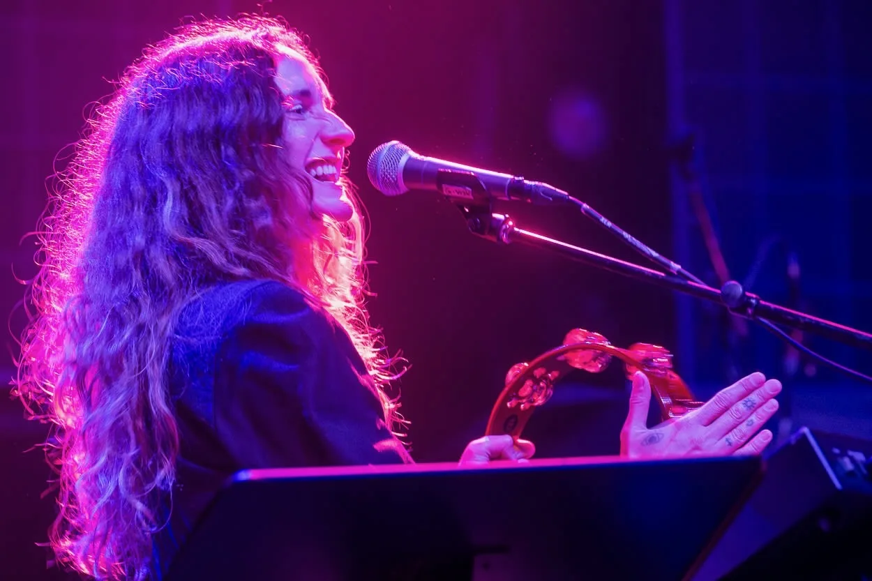 Close-up of Lacey Guthrie singing into a mic with her hand raised, long curly hair lit by purple stage light.