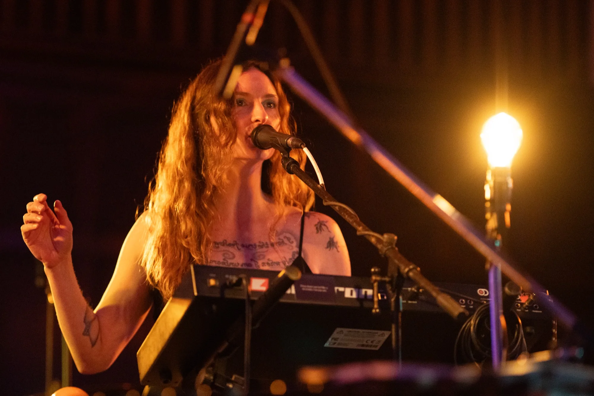Close-up of Lacey Guthrie singing into a microphone at a keyboard, lit by a warm stage light.
